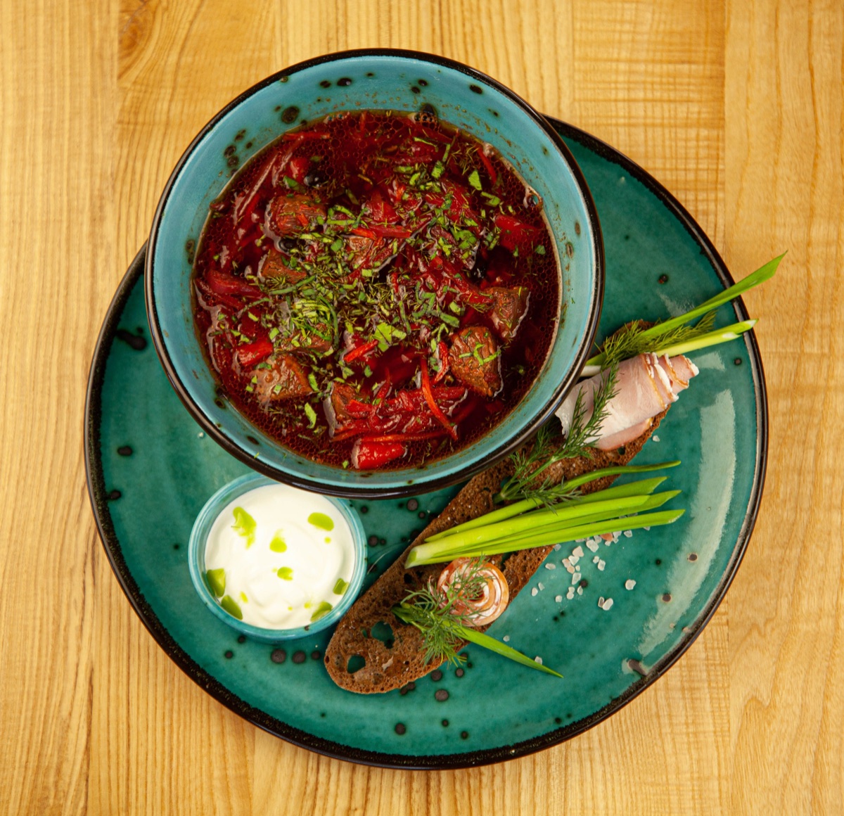 Домашний борщ с мясом в бирюзовой миске, рядом сметана, ржаной хлеб и зелёный лук
